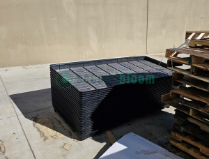 A tall stack of black plastic Botanicare trays stored outdoors against a beige wall.