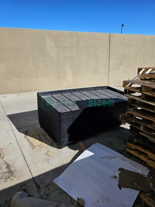 A tall stack of black plastic Botanicare trays stored outdoors against a beige wall.