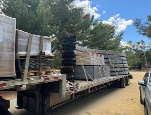 Flatbed truck loaded with disassembled Harnois Ovaltech greenhouse components including metal framework, packaged materials, and structural elements