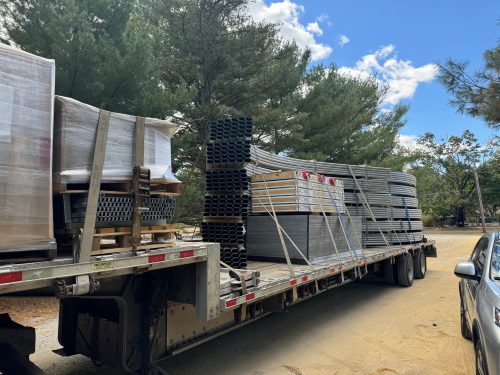 Flatbed truck loaded with disassembled Harnois Ovaltech greenhouse components including metal framework, packaged materials, and structural elements