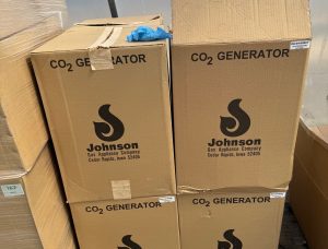Four boxed Johnson Gas Appliance Company CO2 generators stacked inside a greenhouse, with cardboard packaging showing the manufacturer's logo