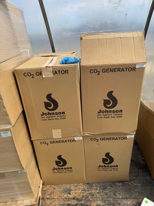 Four boxed Johnson Gas Appliance Company CO2 generators stacked inside a greenhouse, with cardboard packaging showing the manufacturer's logo