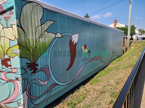 Side view of blue shipping container farm with painted mural of green plants, pink water waves, and #eatlocal hashtag along residential street