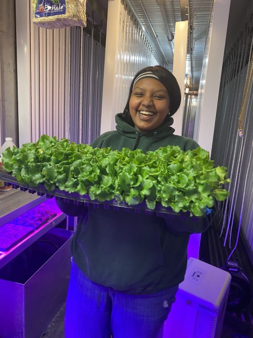 Smiling woman in green hoodie holding large tray of fresh leafy green lettuce inside hydroponic farm