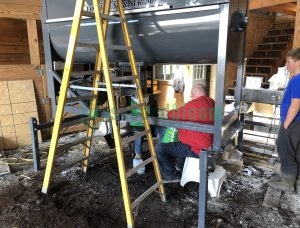 Industrial soil mixer machine with large gray hopper, yellow ladder access, and control panel being inspected by technicians in a warehouse setting